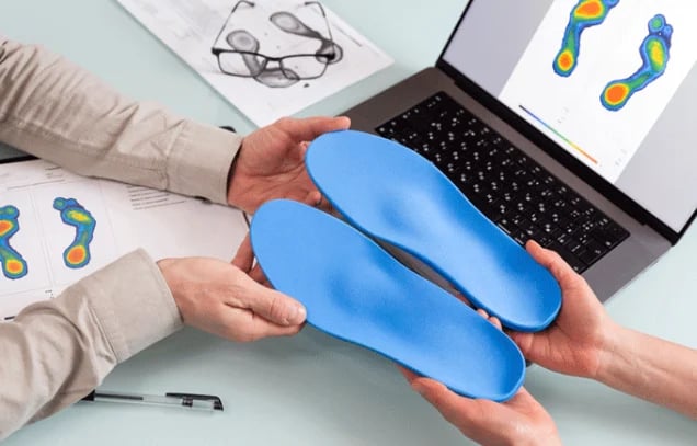 Healthcare professional holding blue orthotic shoe insoles at desk with laptop and thermal foot scan images